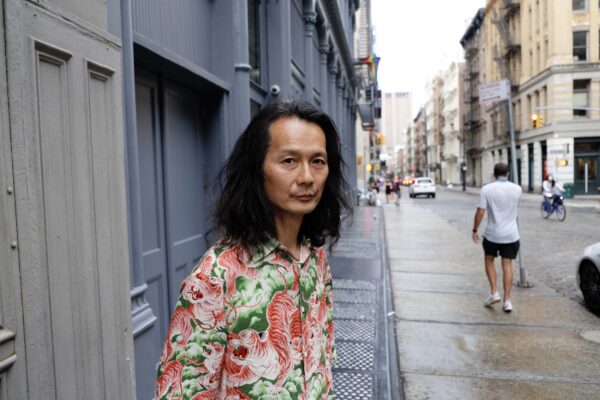 Person with long hair and a colorful shirt stands on a sidewalk in a lively city with buildings and pedestrians.
