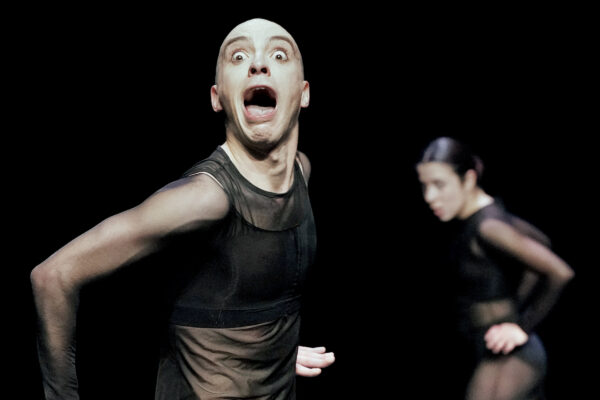 Two actors in dark clothing pose theatrically: one with an open mouth and the other blurred in the background."