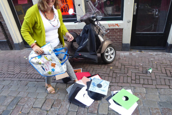 Smiling woman in green cardigan and colourful necklace holds a bag where vinyl records have just fallen out of, lying on the floor.
