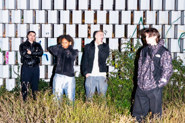 Four people stand in front of a patterned white-and-gray block wall, surrounded by overgrown grass.