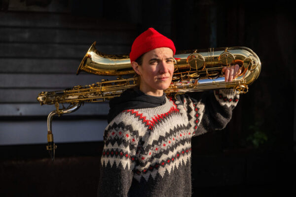 Person with a red bonnet and a gold-coluored saxophone over their shoulder, wearing a patterned sweater.