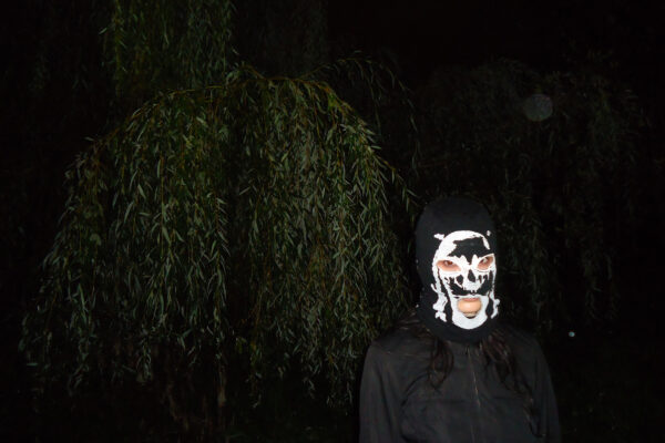 Person wears a black mask with a white skull print, standing before dark trees with drooping branches.