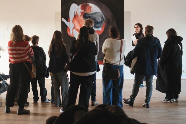 A group of people views an abstract painting with warm colours in a brightly lit gallery space.