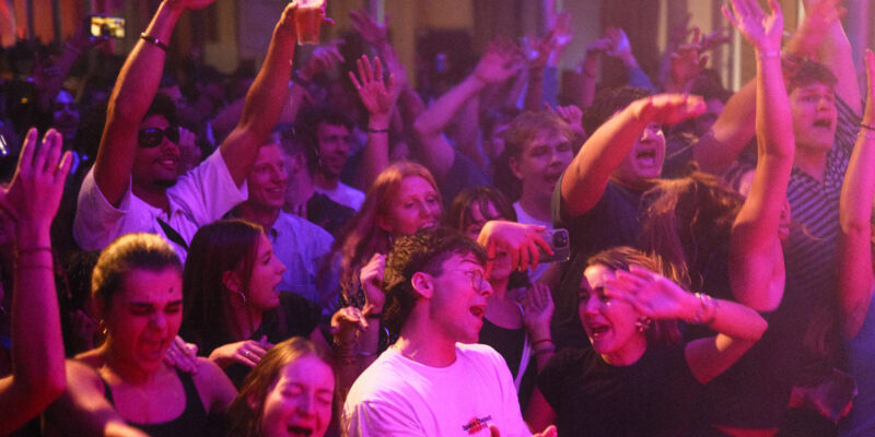 Een energieke menigte jonge volwassenen die enthousiast samen dansen op een feest met fel paarse en roze podiumverlichting, handen omhoog in vreugdevolle viering

English: An energetic crowd of young adults dancing enthusiastically together at a party with vibrant purple and pink stage lighting, hands raised in joyful celebration