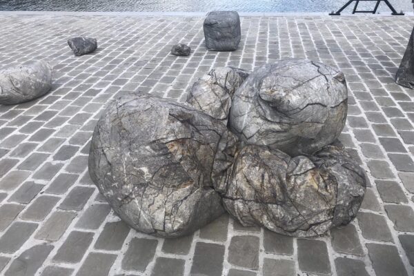 Steenachtige vormen ter grootte van een mens liggen op straat, op kasseien naast het water. 

Rock-like shapes the size of a person lie on the street, on a cobbled are next to a bassin of water.