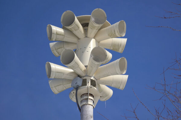 Buitenwaarschuwingssirenes met meerdere hoornluidsprekers op een paal, van onderaf gezien tegen een heldere blauwe lucht.

Outdoor warning siren with multiple horn speakers mounted on a pole, viewed from below against a clear blue sky and bare tree branches.