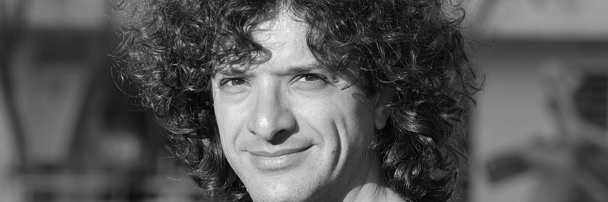 Zwart-wit portret van een persoon met dik krullend haar in donker t-shirt, subtiel glimlachend, met zacht vervaagde binnenruimte op de achtergrond.

Black and white close-up portrait of a person with thick curly hair wearing a dark t-shirt, smiling subtly, with softly blurred indoor background