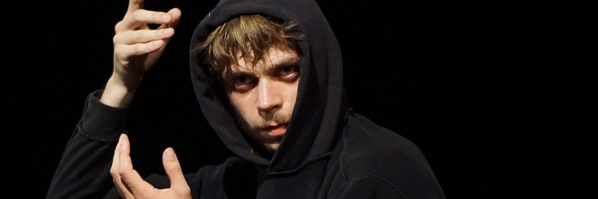 Persoon in een zwarte hoodie die optreedt op een podium, gefotografeerd tijdens een beweging met uitgestrekte handen in zwak licht.