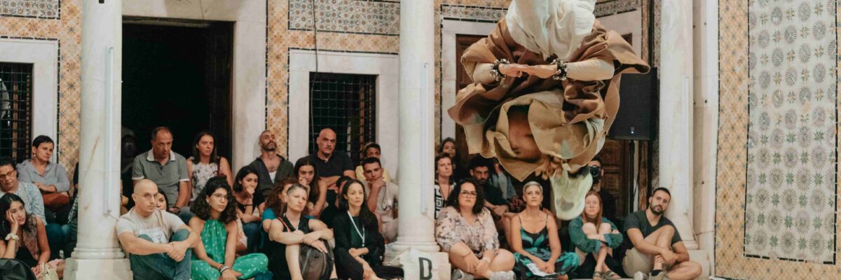 Een danser in een beige, vloeiende kostuum springt in de lucht in een weelderig betegelde zaal met toeschouwers.

A dancer in a beige, flowing costume jumps mid-air in an ornate, tiled hall with seated spectators.
