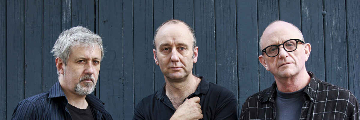 Drie mannen staand tegen donkere houten muur: links met grijs haar in gestreept overhemd, midden in zwart polo met gekruiste armen, rechts met bril in geruit overhemd. 

Three men standing against dark wooden wall: left with grey hair in striped shirt, center in black polo with arms crossed, right with glasses in checkered shirt.