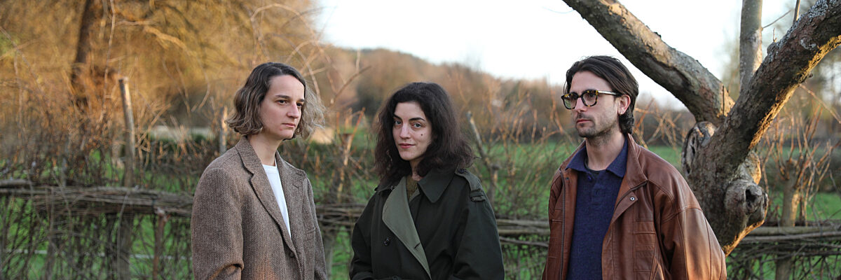 Drie personen staand in veld met houten hek en kale bomen: links in tweed jas, midden in donkere jas, rechts in bruine leren jas met bril. 

Three people standing in field with wooden fence and bare trees: left in tweed coat, center in dark coat, right in brown leather jacket with glasses.