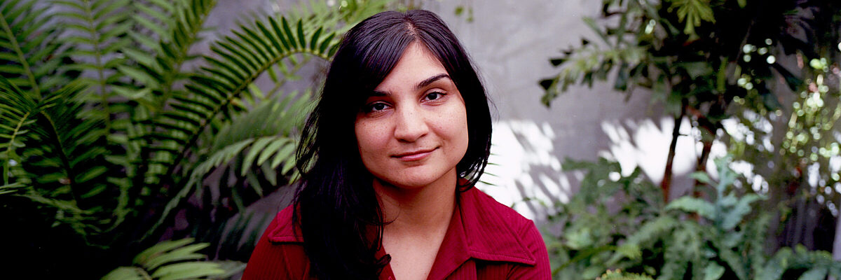 Een persoon met lang donker haar in een rode blouse, zittend tussen weelderige groene varens en hangplanten tegen een betonnen muur

English: A person with long dark hair wearing a red shirt, sitting among lush green ferns and hanging plants against a concrete wall