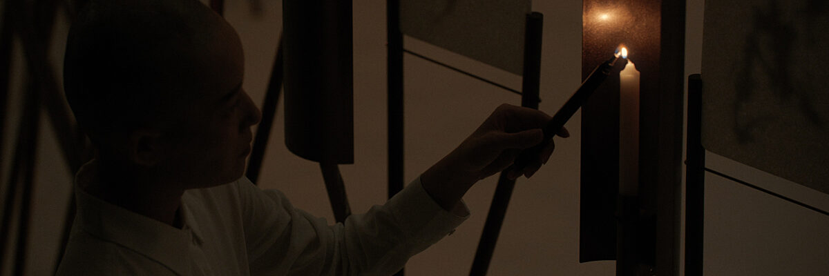 Iemand in een donkere ruimte steekt een kaars aan die half omringd is door een metalen plaat. 

Person in a dark room lighting a candle standing on a metal holder, half encircled by a metal panel.