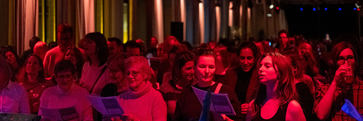 Een grote groep mensen staat in een warm verlichte ruimte, met liedteksten in de hand, en zingt samen; sommigen dragen een bril en de verlichting is gedempt met rode en blauwe tinten.
A large group of people, standing in a warmly lit room, hold song sheets and sing together; some use glasses, and lighting is low with red and blue tones.