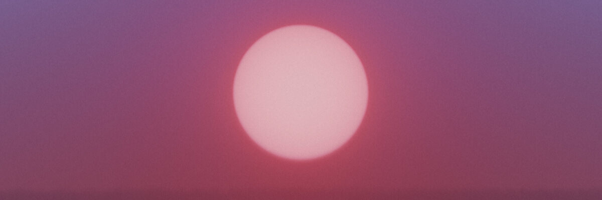 Purple and pink sky at sunset with a brightly lit sun above dark trees.