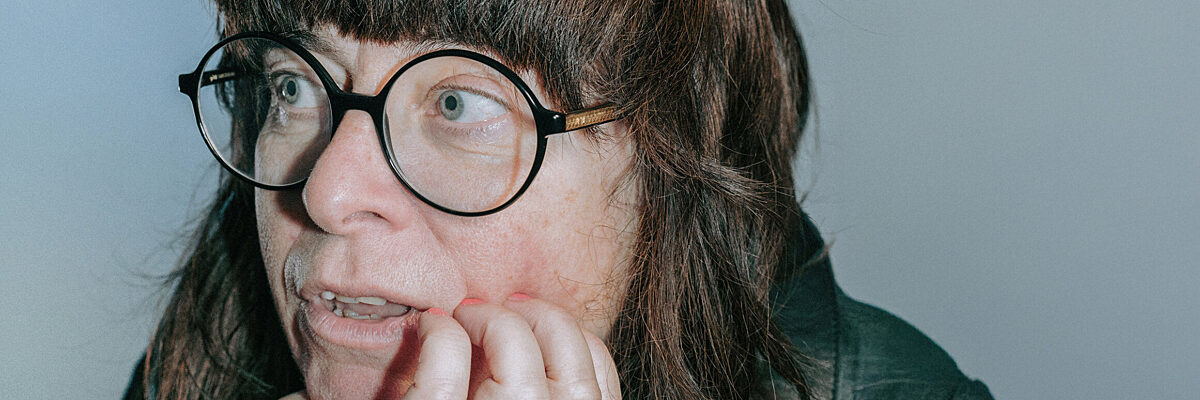 A person with long dark hair and round glasses rests their chin on their hand, looking thoughtful against a plain light background.