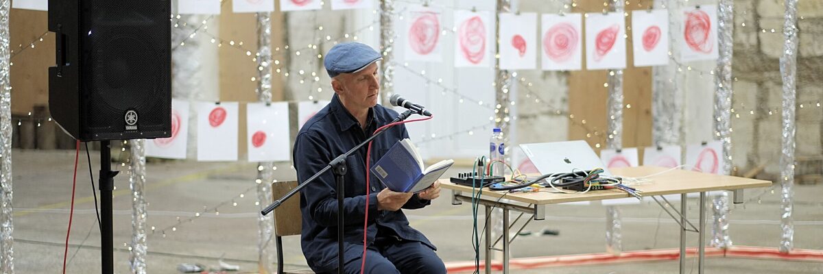 Een man zit op een stoel in een grote, lichte ruimte en leest voor uit een boek. Naast de man staat een luidspreker en een tafel met audiomateriaal en een laptop. Achteraan de ruimte hangen tekeningen van rode cirkels aan kabels.
A man sits on a chair in a big, bright room and reads from a book, sitting between a large speaker and a table with audio equipment and a computer on it. In the background, dozens of drawings of red circles hang throughout the room.