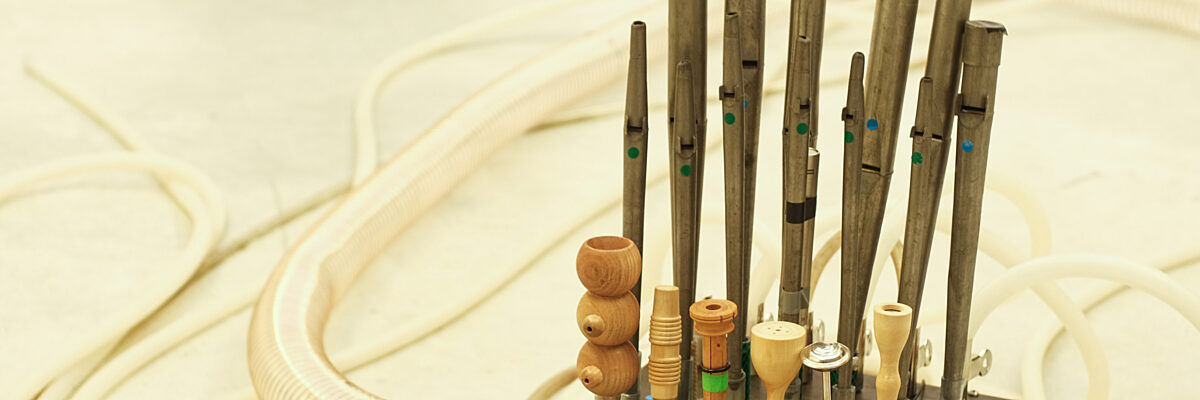 Muziekinstrument met rechtopstaande fluiten in hout en metaal verbonden met een zwarte box waar een witte luchtslang aan hangt. 

Musical instrument with flutes in wood and metal, all standing on a black metal box that is attached to a large white air hose.