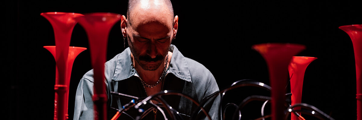 Man bespeelt geconcentreerd een instrument met knoppen op een horizontaal vlak omringd door rechtopstaande trompetten. 

Man in full concentration plays an instrument with buttons on a horizontal area surrounded by trumpets standing up.