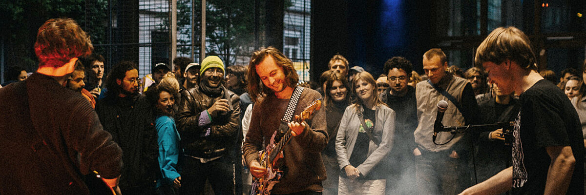 Muzikanten treden live op met gitaren en synthesizers terwijl een glimlachend publiek zich verzamelt in een rokerige, sober verlichte zaal.

Musicians perform live with guitars and synths as a smiling crowd gathers in a smoky, dimly lit indoor venue.