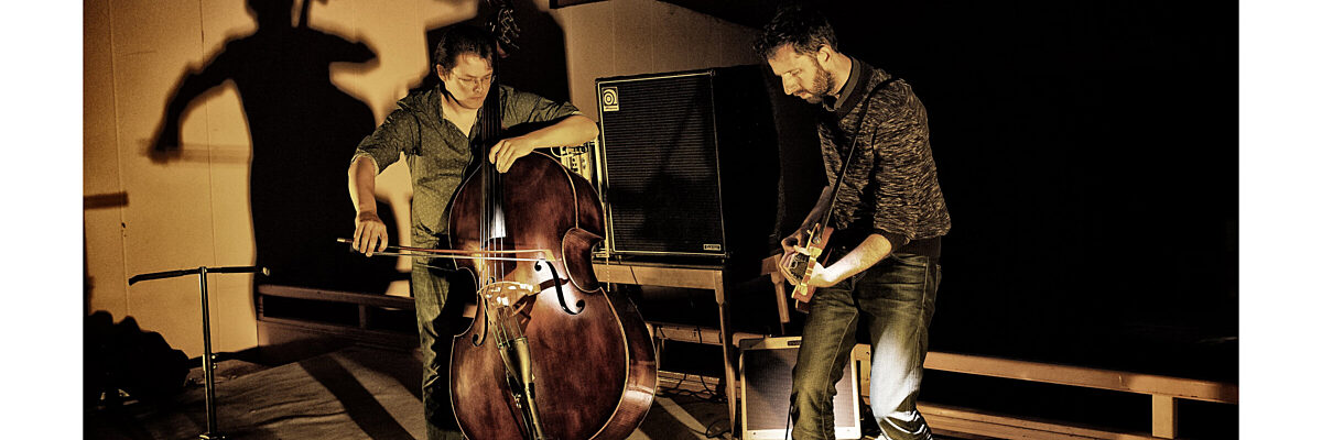 Two musicians perform on stage: one plays a double bass, the other an electric guitar. Shadows and warm lighting create a dynamic jazz atmosphere.