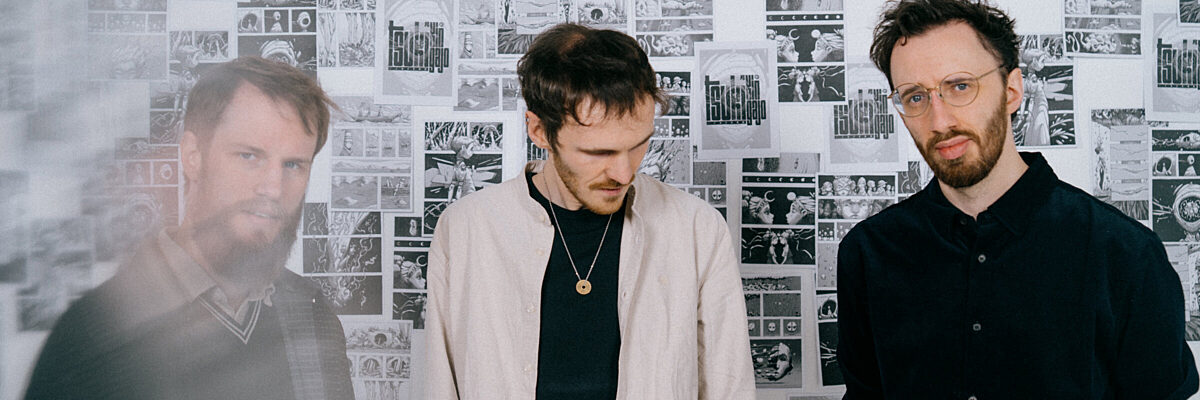Three men stand in a room covered with intricate black-and-white comic-style artwork, one wearing glasses and a black shirt, the other in a light jacket, the third a black sweater.