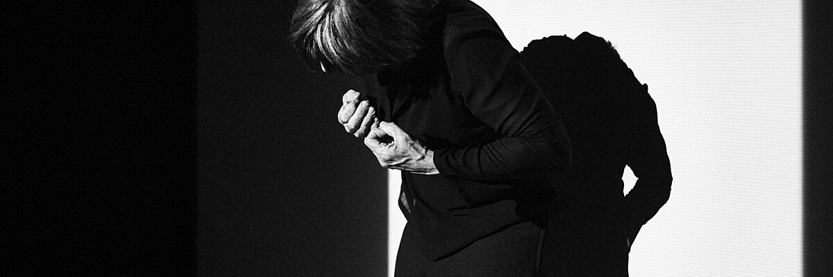 Een danser staat voorovergeboven met de rug naar een muur, het gezicht onherkenbaar, de handen in vuisten voor de borst geklemd.  

A dancer stands with the back against a wall, bending forward, face hidden, hands in clenched fists in front of the person.