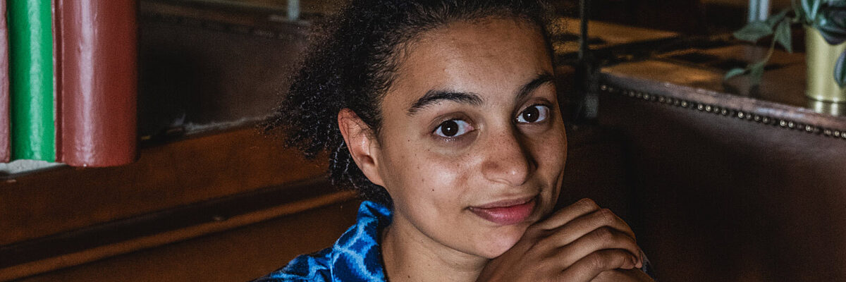 Vrouw zit in een diner in een blauw kleed, haar ellebogen rustend op de tafel, handen ondersteunen de kin, en lacht naar de camera.

Woman sitting in a diner wearing a blue patterned dress, elbows resting on the table, hands supporting her head, smiling into the camera.