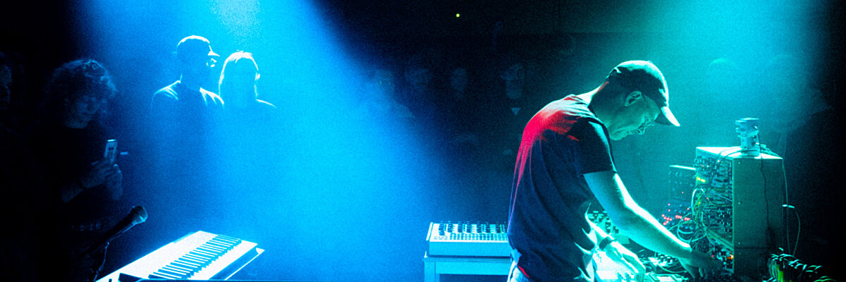 Ruim zijwaarts beeld van een man in een clubsetting die een analogue synth bespeelt, belicht door groene en blauwe spots. 

Side shot of man in club setting performing on an analogue synth, lit by green and blue spots.