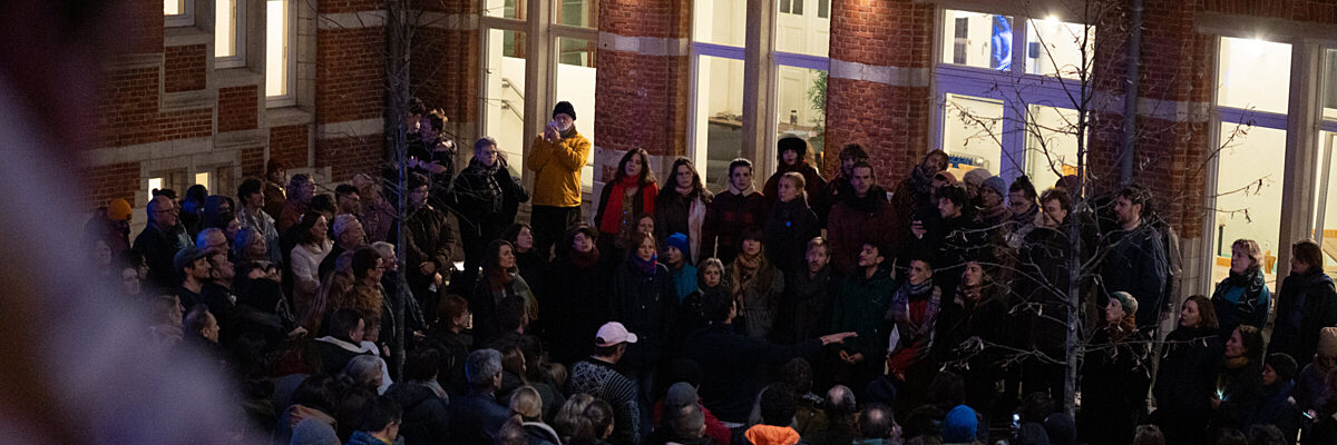 Een grote menigte in winterjassen verzamelt 's nachts buiten een bakstenen gebouw met verlichte ramen, gericht naar het gebouw onder kale bomen.