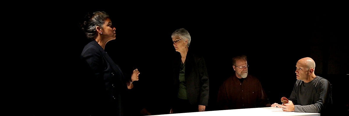 Vijf volwassenen zitten of staan rond een verlichte tafel in een donkere kamer. Eén vrouw spreekt, de anderen luisteren. Een man houdt een dennenappel vast.
Five adults sit or stand around a lit table in a dark room. One woman speaks firmly, while others listen seriously. One man holds a pinecone.