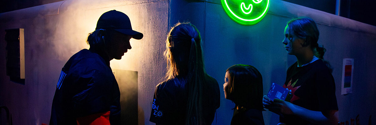 Vier mensen staan in een donkere ruimte met een neon groen berenhoofd-teken op de muur achter hen. Eén draagt een pet en een rode armband.