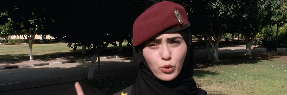 Vrouw in legeruniform met Palestijnse badges en een rode baret staat in een park en houdt haar rechter wijsvinger omhoog.

Woman in army uniform with Palestine badges and a red baret stands in a park holding up her right index finger.
