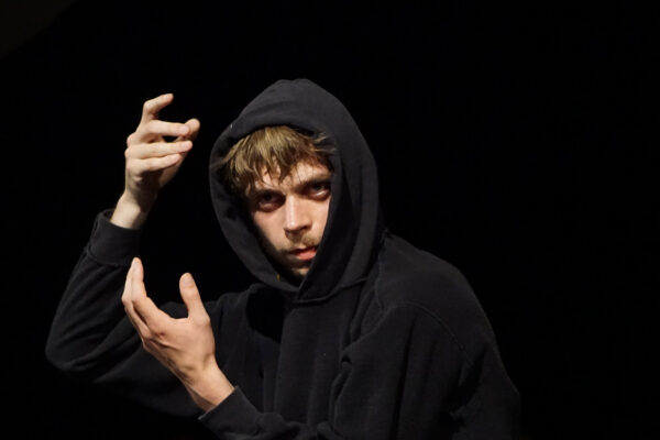 Persoon in een zwarte hoodie die optreedt op een podium, gefotografeerd tijdens een beweging met uitgestrekte handen in zwak licht.