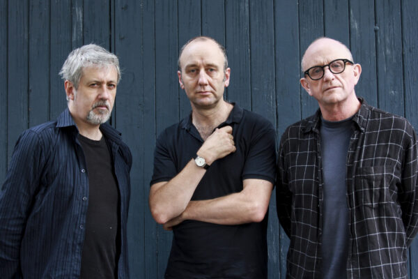 Drie mannen staand tegen donkere houten muur: links met grijs haar in gestreept overhemd, midden in zwart polo met gekruiste armen, rechts met bril in geruit overhemd. 

Three men standing against dark wooden wall: left with grey hair in striped shirt, center in black polo with arms crossed, right with glasses in checkered shirt.