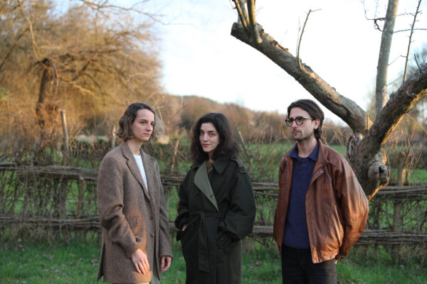 Drie personen staand in veld met houten hek en kale bomen: links in tweed jas, midden in donkere jas, rechts in bruine leren jas met bril. 

Three people standing in field with wooden fence and bare trees: left in tweed coat, center in dark coat, right in brown leather jacket with glasses.