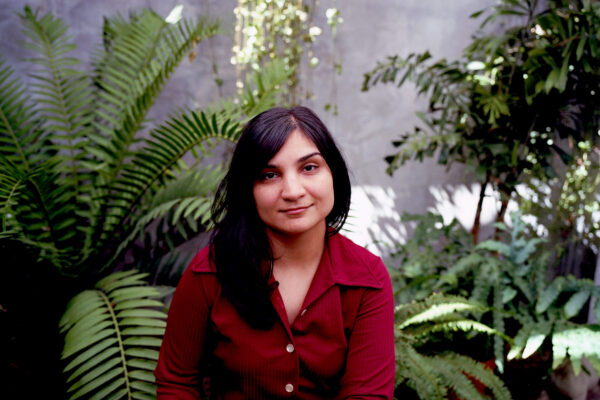 Een persoon met lang donker haar in een rode blouse, zittend tussen weelderige groene varens en hangplanten tegen een betonnen muur

English: A person with long dark hair wearing a red shirt, sitting among lush green ferns and hanging plants against a concrete wall