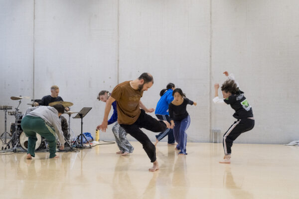Groep artiesten danst in een studio, terwijl een drummer op de achtergrond speelt, in een levendige en artistieke repetitie.

Group of performers dances dynamically in a studio, while a drummer plays in the background, creating a lively and artistic rehearsal scene.