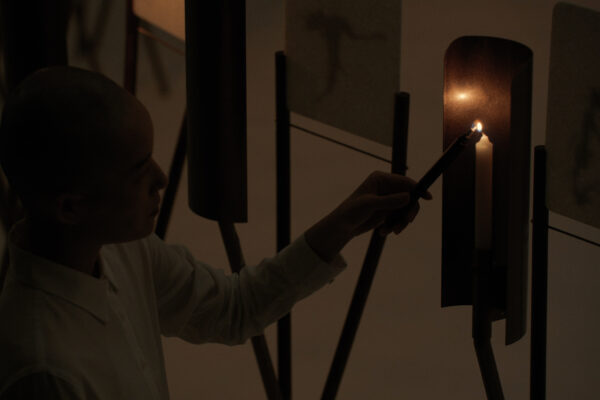 Iemand in een donkere ruimte steekt een kaars aan die half omringd is door een metalen plaat. 

Person in a dark room lighting a candle standing on a metal holder, half encircled by a metal panel.