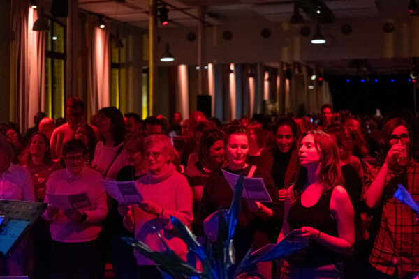 Een grote groep mensen staat in een warm verlichte ruimte, met liedteksten in de hand, en zingt samen; sommigen dragen een bril en de verlichting is gedempt met rode en blauwe tinten.

A large group of people, standing in a warmly lit room, hold song sheets and sing together; some use glasses, and lighting is low with red and blue tones.