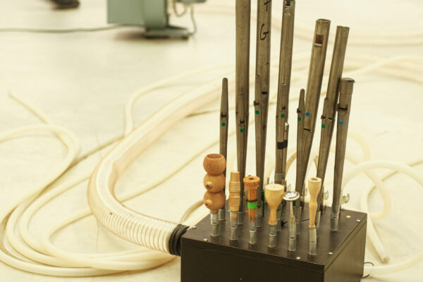 Muziekinstrument met rechtopstaande fluiten in hout en metaal verbonden met een zwarte box waar een witte luchtslang aan hangt. 

Musical instrument with flutes in wood and metal, all standing on a black metal box that is attached to a large white air hose.