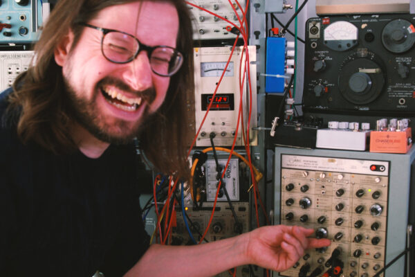 Een man met lang haar, een bril en een baard glimlacht breed terwijl hij aan een knopje draait op een paneel met vintage audiomateriaal. Meerdere kabels verbinden verschillende toestellen in de achtergrond, wat de suggestie wekt dat het om een experimentele audio-setup gaat. 

A man with long hair, glasses, and a beard smiles widely while adjusting a knob on a panel of vintage electronic audio equipment. Multiple wires connect various devices in the background, suggesting an experimental or sound engineering setup.
