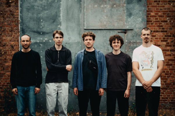 Vijf mannen staan in een rij voor een industriële bakstenen en metalen achtergrond, poserend voor een groepsfoto in casual kleding.