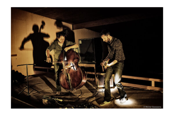 Two musicians perform on stage: one plays a double bass, the other an electric guitar. Shadows and warm lighting create a dynamic jazz atmosphere.
