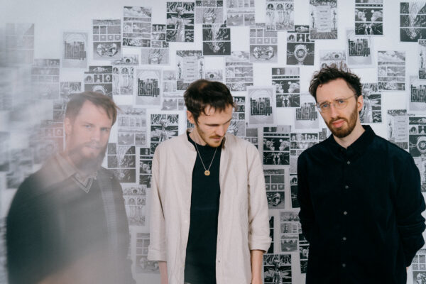 Three men stand in a room covered with intricate black-and-white comic-style artwork, one wearing glasses and a black shirt, the other in a light jacket, the third a black sweater.
