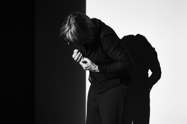 Een danser staat voorovergeboven met de rug naar een muur, het gezicht onherkenbaar, de handen in vuisten voor de borst geklemd.  

A dancer stands with the back against a wall, bending forward, face hidden, hands in clenched fists in front of the person.