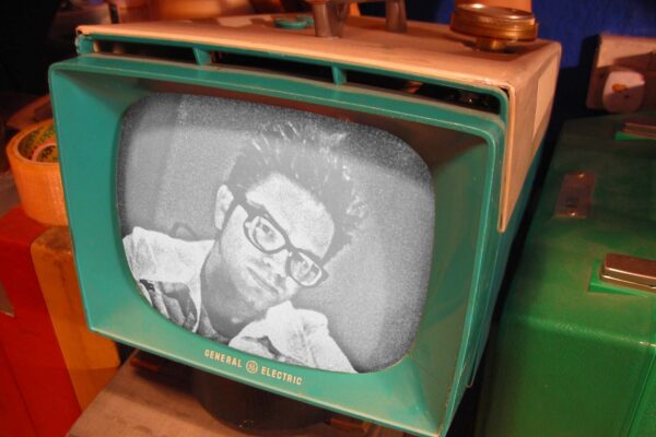 Een vintage groene en beige General Electric-televisie toont een zwart-wit close-up van iemand met een bril. Andere onscherpe objecten staan rond de TV.