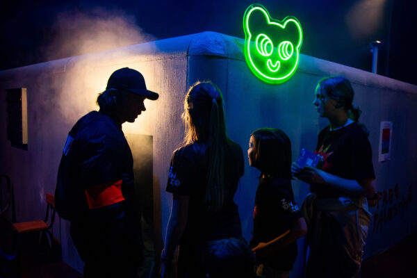 Vier mensen staan in een donkere ruimte met een neon groen berenhoofd-teken op de muur achter hen. Eén draagt een pet en een rode armband.