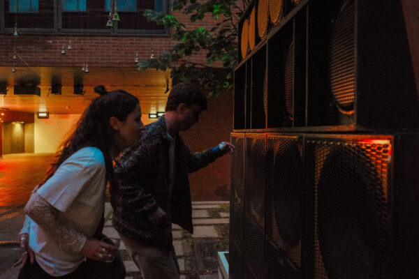 Een vrouw en een man staan te dansen vlak voor een soundsystem bestaande uit meerdere luidsprekers die naast elkaar en op elkaar gestapeld zijn, als een wall of sound. 

A woman and a man are dancing in front of a soundsystem, consisting of several speakers stacked next to and on top of each other, creating a wall of sound.