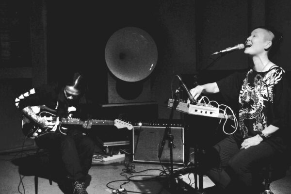 Twee muzikanten treden op in een gedimde ruimte: één speelt elektrische gitaar, de andere zingt in een microfoon met een klein elektronisch apparaat.